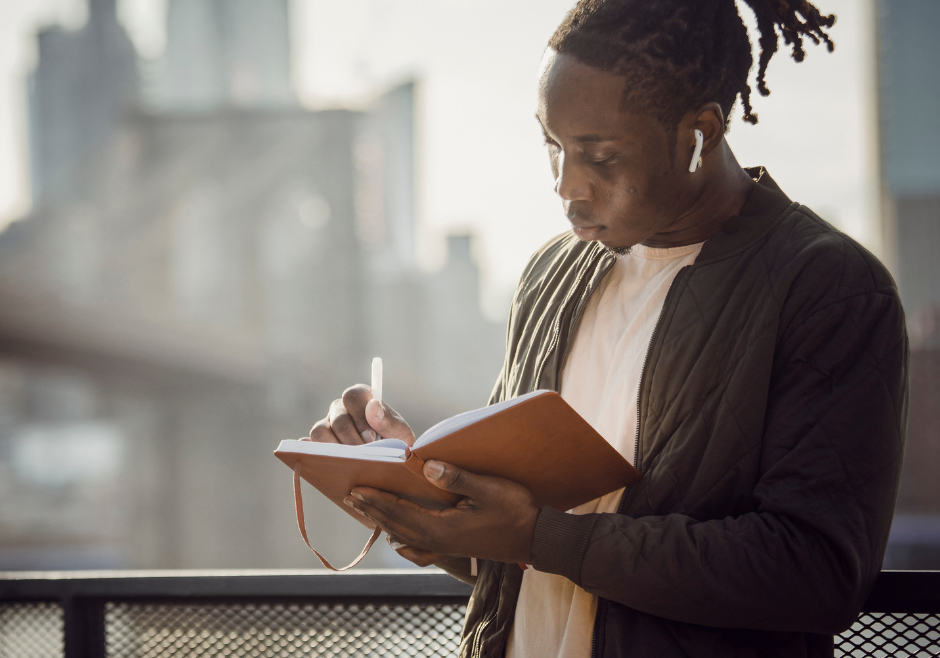 A person is stood, looking at their notebook in deep though as they write a piece of creative non-fiction